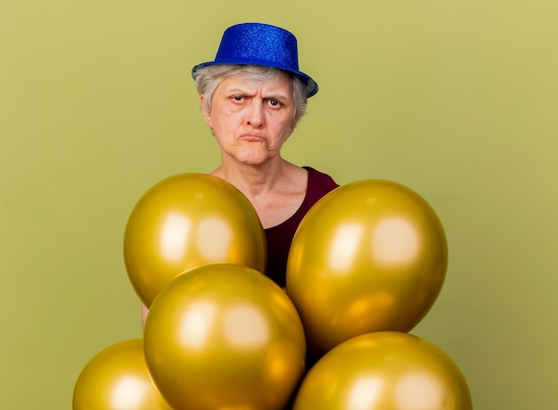 Anciana decepcionada con sombrero de fiesta se encuentra con globos de helio aislados en la pared verde oliva con espacio de copia