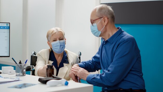 Anciana con discapacidad crónica y su esposo en la cita, asistiendo a la visita de control con un especialista médico. Paciente usuario de silla de ruedas consultando durante la pandemia de covid 19.