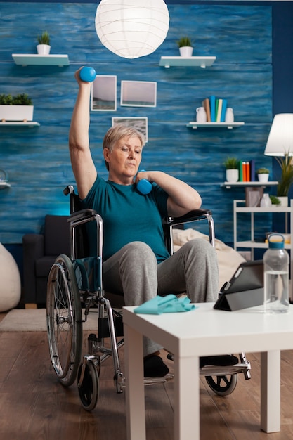 Anciana discapacitada en silla de ruedas levantando la resistencia de los músculos de entrenamiento del brazo con mancuernas