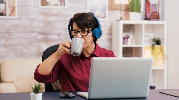 Anciana disfrutando de una taza de café mientras trabaja en la computadora portátil con auriculares en la cabeza. Anciano con tableta en el fondo.