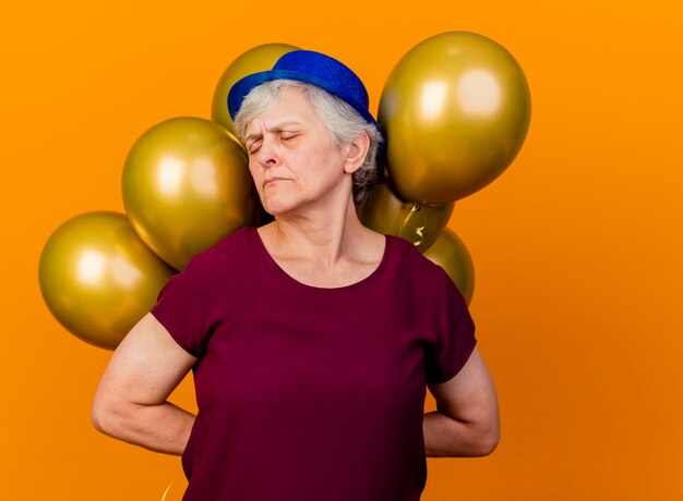 Anciana disgustada con gorro de fiesta sostiene globos de helio detrás con los ojos cerrados en naranja