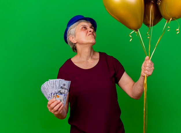Anciana disgustada con gorro de fiesta tiene dinero y mira globos de helio en verde