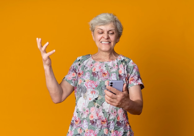 Anciana disgustada levanta la mano sosteniendo y mirando el teléfono aislado en la pared naranja