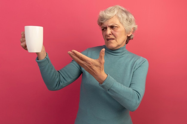 Anciana disgustada vistiendo suéter de cuello alto azul sosteniendo mirando y apuntando con la mano a la taza de té