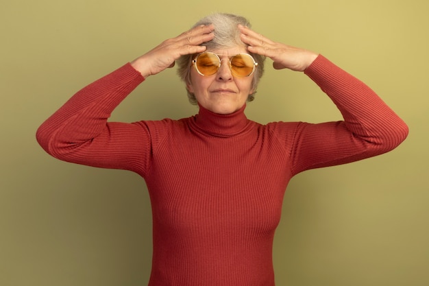 Anciana dolorida con suéter de cuello alto rojo y gafas de sol manteniendo las manos en la cabeza con los ojos cerrados aislados en la pared verde oliva