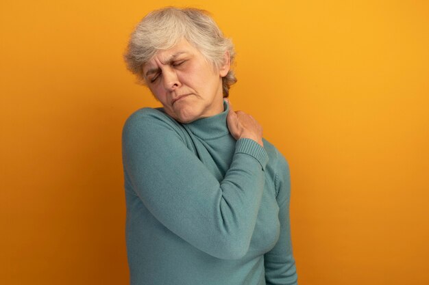 Anciana dolorida vistiendo suéter de cuello alto azul poniendo la mano sobre el hombro con los ojos cerrados