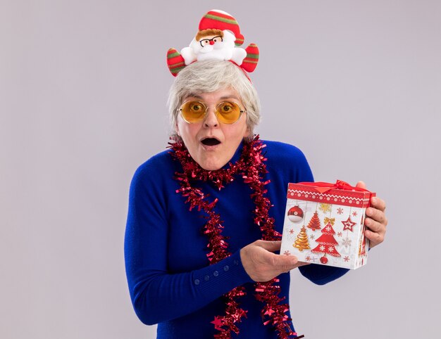 Anciana emocionada en gafas de sol con diadema de santa y guirnalda alrededor del cuello tiene caja de regalo de Navidad aislada en la pared blanca con espacio de copia