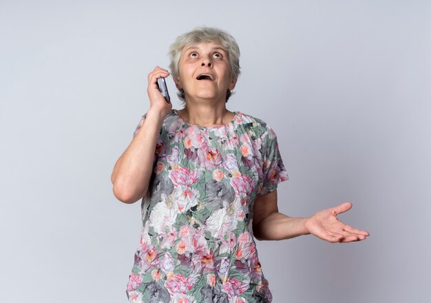 Anciana emocionada habla por teléfono y mira hacia arriba aislado en la pared blanca