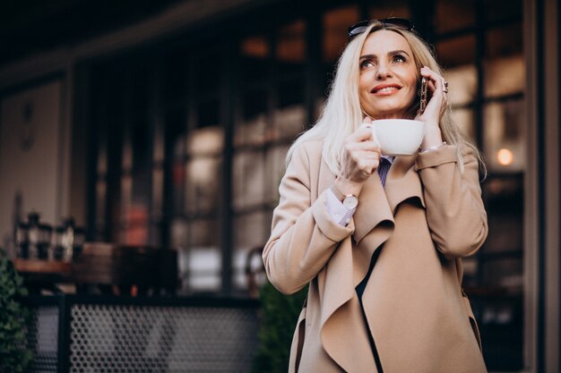 Anciana empresaria tomando café fuera de la cafetería y hablando por teléfono