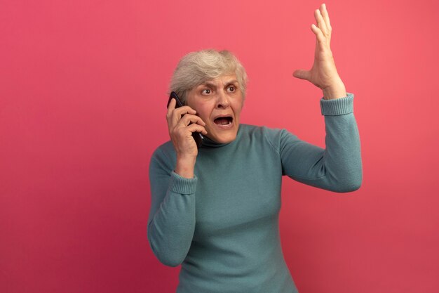 Anciana enojada vistiendo suéter de cuello alto azul hablando por teléfono mirando al lado levantando la mano aislada en la pared rosa con espacio de copia