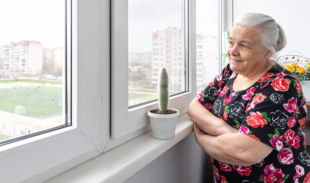 Una anciana está de pie junto a la ventana y mira a lo lejos.