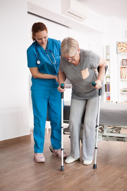 Anciana en hogar de ancianos caminando con muletas con la ayuda de una enfermera.