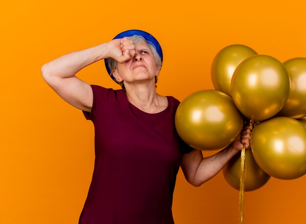 Foto gratuita anciana insatisfecha con sombrero de fiesta sostiene globos de helio y pone el puño en el ojo mirando hacia arriba aislado en la pared naranja con espacio de copia