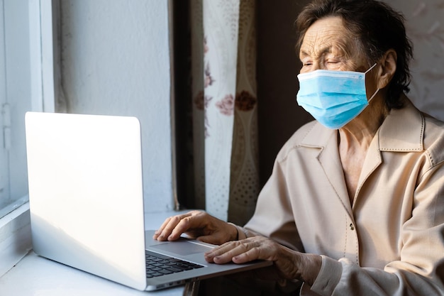 una anciana con una máscara cerca de la ventana con una computadora portátil.