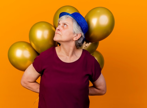 Anciana molesta con gorro de fiesta sostiene globos de helio detrás mirando hacia arriba en naranja