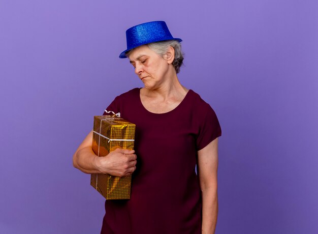 Anciana molesta con gorro de fiesta sostiene y mira caja de regalo aislada en pared púrpura con espacio de copia