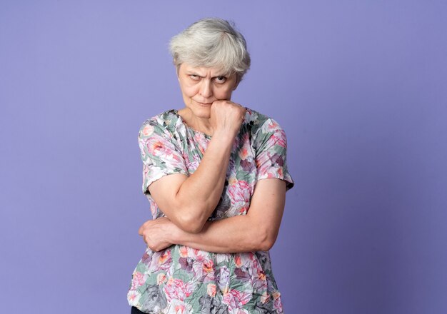 Anciana molesta pone la mano en la barbilla mirando hacia adelante aislado en la pared púrpura