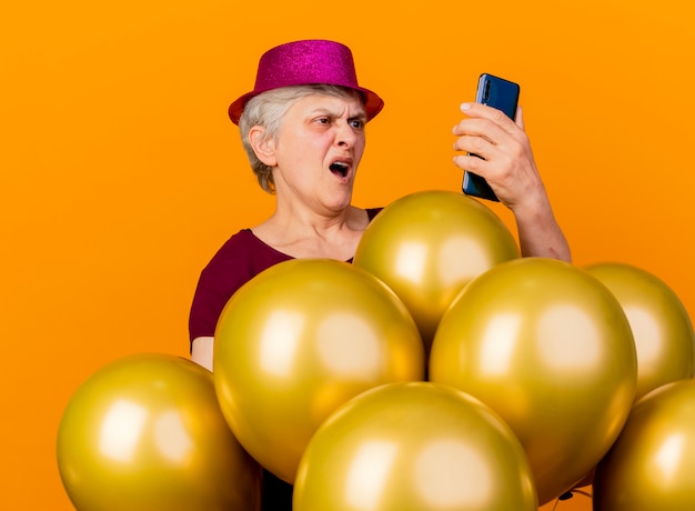 Anciana molesta con sombrero de fiesta se encuentra con globos de helio mirando el teléfono aislado en la pared naranja