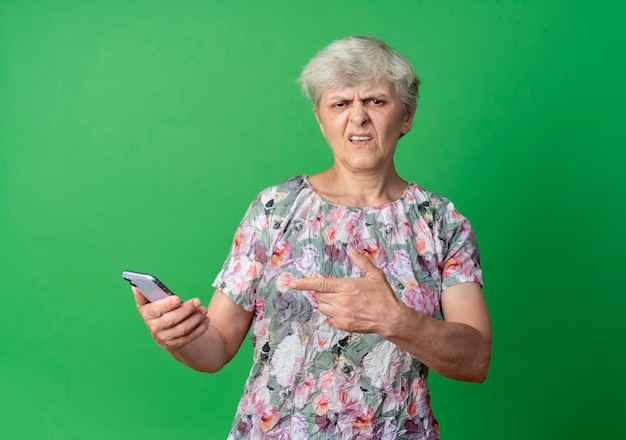 Anciana molesta sostiene y apunta al teléfono aislado en la pared verde
