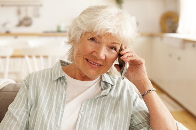 Anciana mujer europea madura en camisa a rayas que tiene una conversación telefónica a través de una aplicación en línea utilizando conexión inalámbrica gratuita a Internet de alta velocidad en casa, mirando con una sonrisa alegre