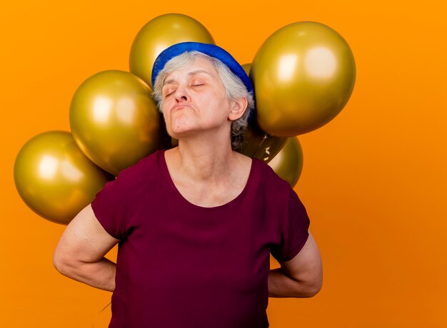 Anciana ofendida con gorro de fiesta sostiene globos de helio detrás con los ojos cerrados en naranja