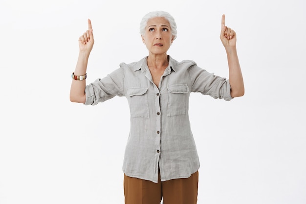Anciana preocupada con cabello gris mirando nerviosa, señalando con el dedo hacia arriba