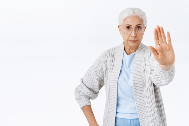 Anciana seria y estricta, la abuela no lo permita, estire el brazo hacia adelante en gesto de parada, rechazo o prohibición, se ve determinada y confiada, no permita que el hijo fume, pared blanca de pie