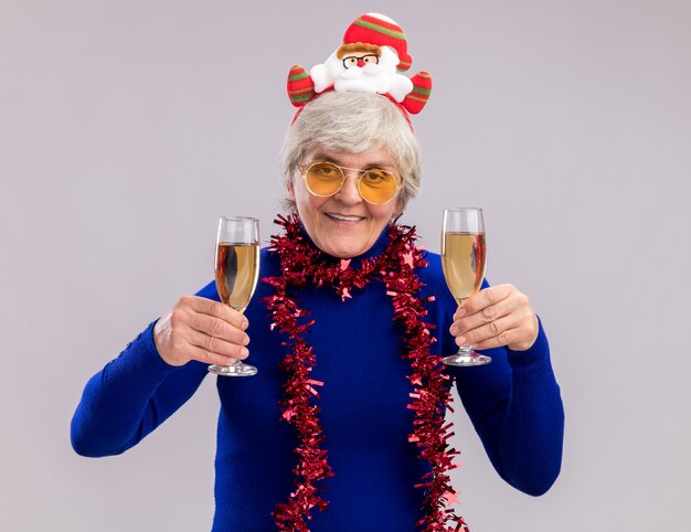 Anciana sonriente en gafas de sol con diadema de santa y guirnalda alrededor del cuello sostiene copas de champán aislado sobre fondo blanco con espacio de copia