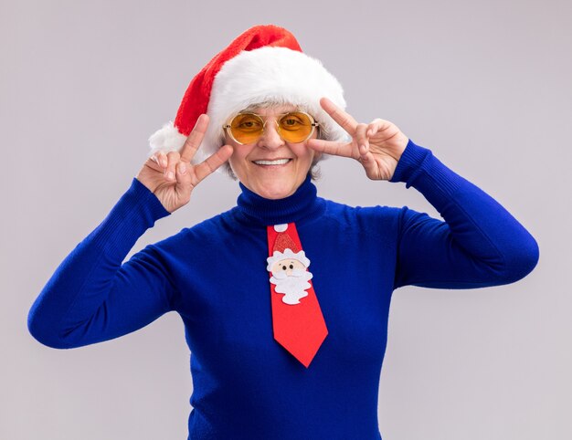 Anciana sonriente en gafas de sol con gorro de Papá Noel y corbata de santa gesticulando el signo de la victoria aislado sobre fondo blanco con espacio de copia