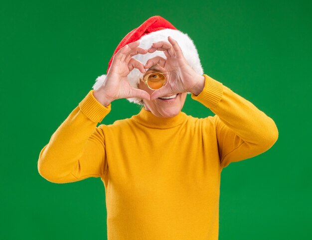 Anciana sonriente en gafas de sol con gorro de Papá Noel a través del signo del corazón