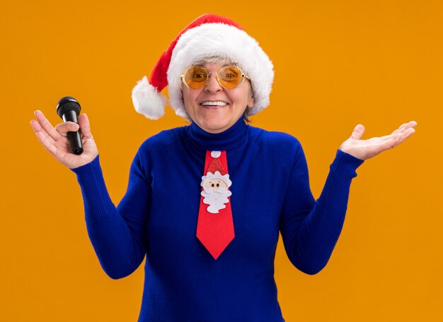 Anciana sonriente en gafas de sol con sombrero de santa y corbata de santa sosteniendo el micrófono y manteniendo la mano abierta aislada en la pared naranja con espacio de copia