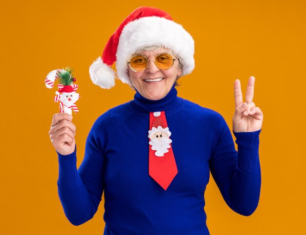 Foto gratuita anciana sonriente en gafas de sol con sombrero de santa y corbata de santa sostiene bastón de caramelo y gestos signo de victoria aislado sobre fondo naranja con espacio de copia