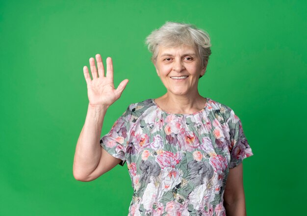 Anciana sonriente levanta la mano aislada en la pared verde