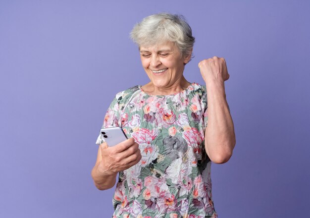 Anciana sonriente levanta el puño mirando el teléfono aislado en la pared púrpura