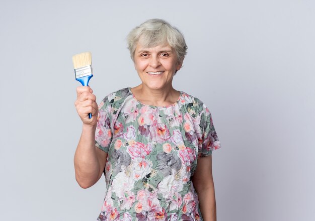 Anciana sonriente sostiene pincel aislado en la pared blanca