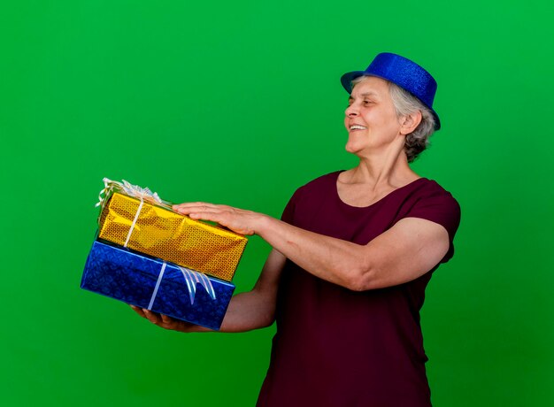 Anciana sonriente vistiendo gorro de fiesta tiene cajas de regalo mirando al lado en verde