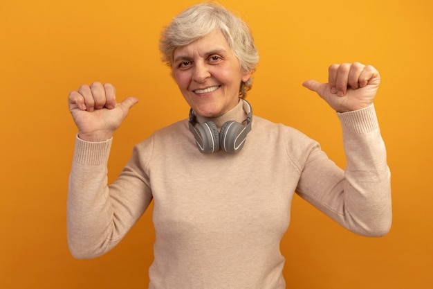 Anciana sonriente vistiendo un suéter de cuello alto cremoso y auriculares alrededor del cuello apuntando hacia atrás