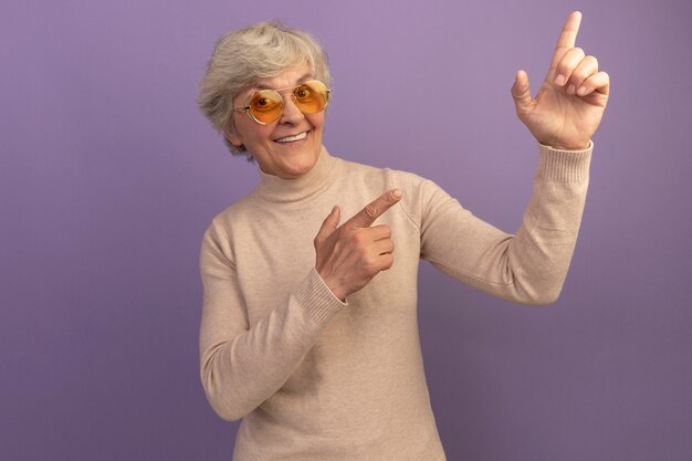 Anciana sonriente vistiendo un suéter de cuello alto cremoso y gafas de sol apuntando hacia arriba