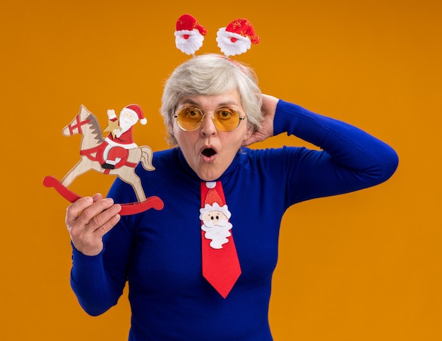 Anciana sorprendida en gafas de sol con diadema de santa y corbata de santa sostiene a santa en la decoración del caballo mecedora aislada sobre fondo naranja con espacio de copia