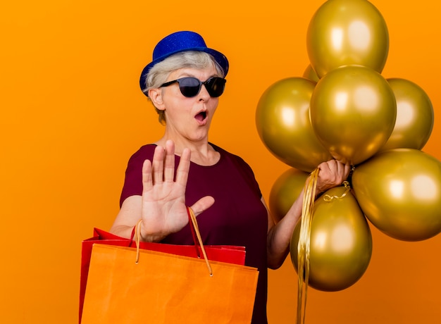 Anciana sorprendida en gafas de sol con sombrero de fiesta sostiene globos de helio y bolsas de papel gesticulando señal de parada aislada en la pared naranja con espacio de copia