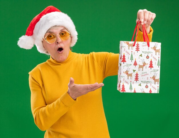 Anciana sorprendida en gafas de sol con sombrero de santa sosteniendo y apuntando a la bolsa de regalo de papel aislada sobre fondo verde con espacio de copia