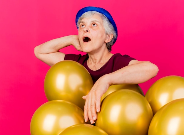 Anciana sorprendida con gorro de fiesta pone la mano en la cabeza detrás de pie con globos de helio