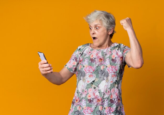 Anciana sorprendida levanta el puño mirando el teléfono aislado en la pared naranja