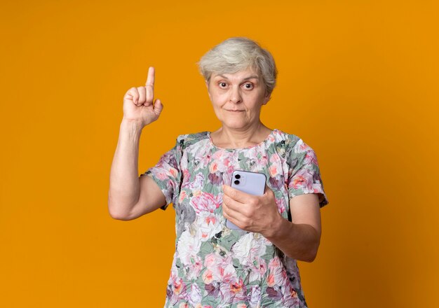 Anciana sorprendida señala sosteniendo el teléfono aislado en la pared naranja