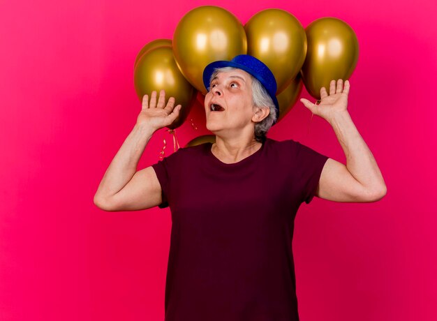 Anciana sorprendida con sombrero de fiesta se encuentra frente a globos de helio con las manos levantadas mirando hacia arriba en rosa