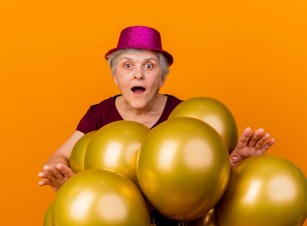 Anciana sorprendida con sombrero de fiesta se encuentra con globos de helio mirando al frente aislado en la pared naranja