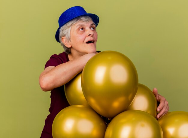 Anciana sorprendida con sombrero de fiesta se encuentra con globos de helio mirando al lado aislado en la pared verde oliva con espacio de copia