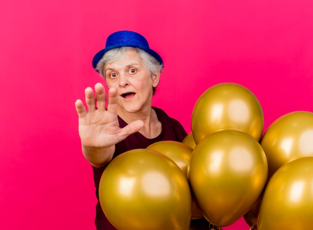 Anciana sorprendida con sombrero de fiesta de pie con globos de helio extendiendo la mano en rosa