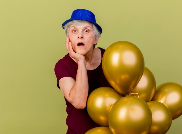 Anciana sorprendida con sombrero de fiesta pone la mano en la barbilla y sostiene globos de helio aislados en la pared verde oliva con espacio de copia