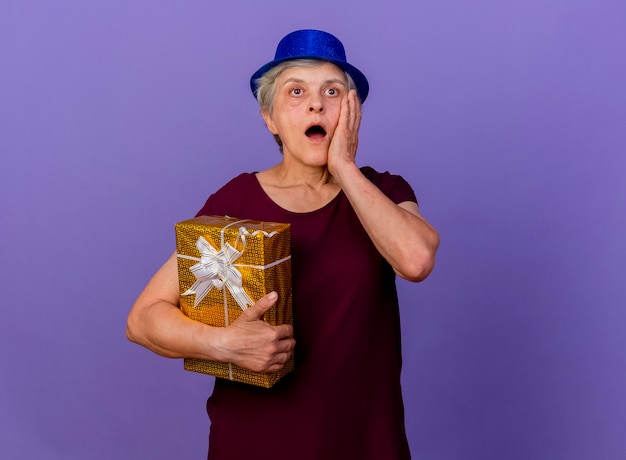 Anciana sorprendida con sombrero de fiesta pone la mano en la cara sosteniendo una caja de regalo aislada en la pared púrpura con espacio de copia